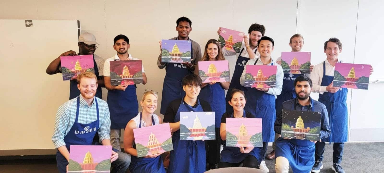 A group of people participating in a DC Paint and Sip event, holding up their paintings in an office.