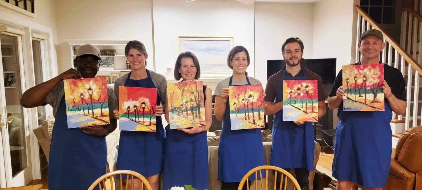 A group of people participating in a DC sip and paint event, holding up paintings in a living room.